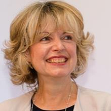 Headshot of a smiling woman with short curly blonde hair, wearing a light jacket over a dark top.