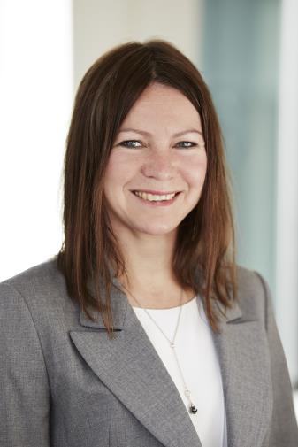 Headshot of a smiling woman with brown hair, wearing a grey jacket and a long necklace.