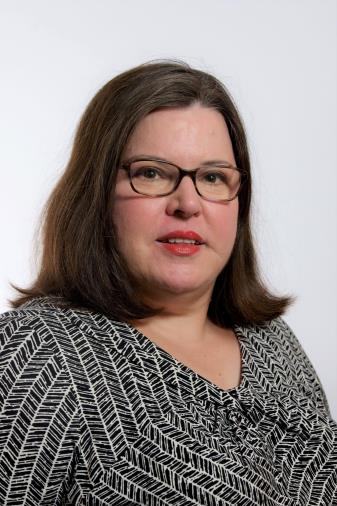 Headshot of a woman with brown hair and glasses, wearing a black and white patterned shirt.