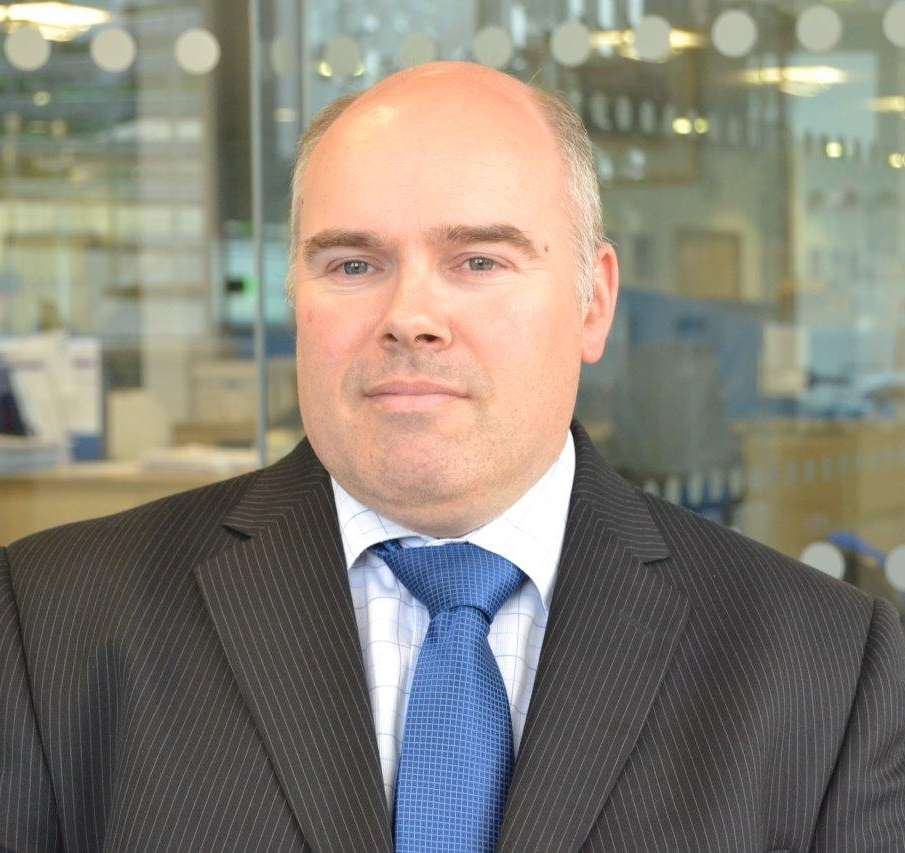 Headshot of a bald man in a dark pinstripe suit and blue patterned tie, looking directly at the camera.
