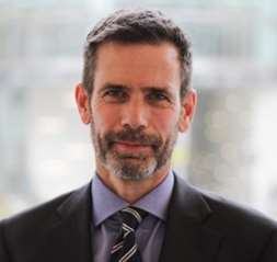 Headshot of a man with short dark hair and a beard, wearing a suit and striped tie.