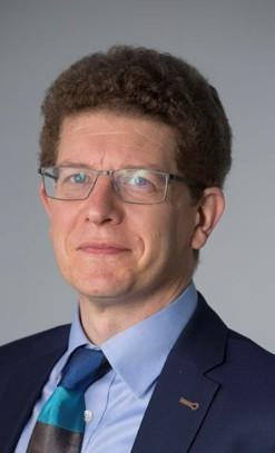 Headshot of a man with curly brown hair and glasses, wearing a blue suit, light blue shirt, and a striped tie.