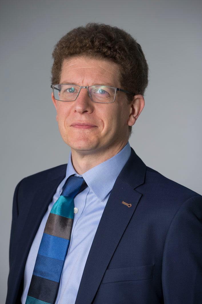 Headshot of a man with glasses, wearing a suit and tie, against a gray background.