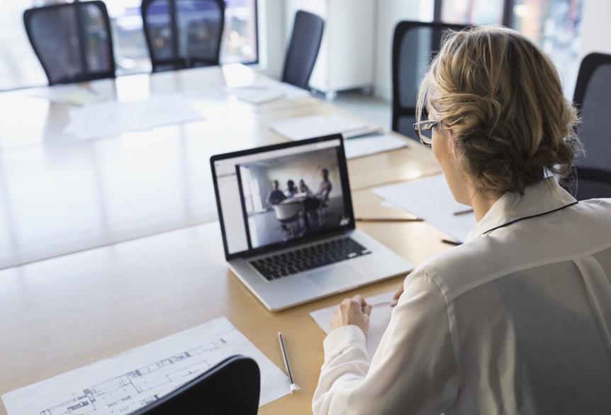 A woman participates in a video conference from a large conference table, viewing other attendees on her laptop.