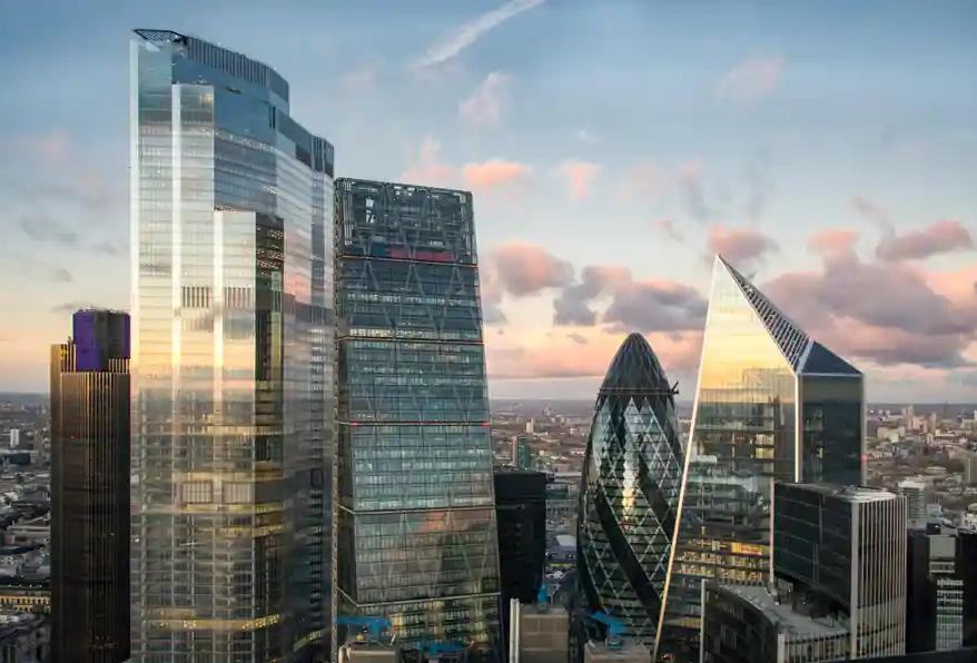 Aerial view of several iconic skyscrapers in the City of London, including the Gherkin, Cheesegrater, and 22 Bishopsgate, under a cloudy sky.